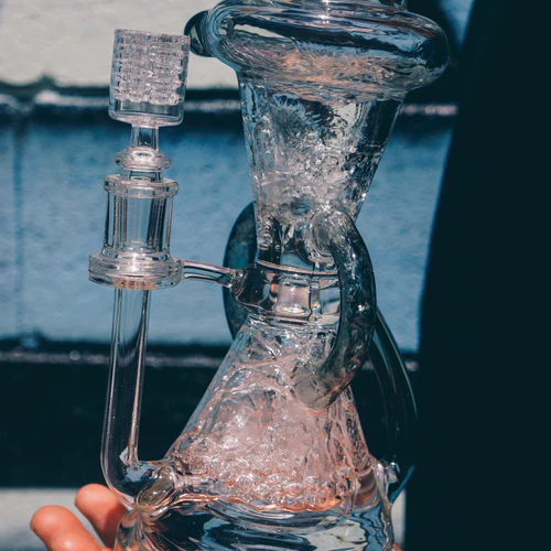 A hand holding a transparent, intricately designed glass water pipe with visible ice cubes inside, set against a blurred blue background.