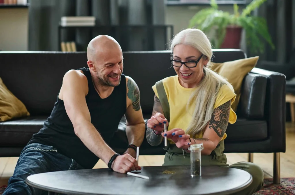 A bald man and a woman with long white hair and glasses, both with tattoos, smiling and looking at a smartphone together at a coffee table.