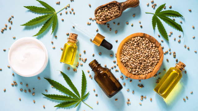 Various hemp products including hemp seeds, oil, and leaves are spread out on a light blue background. Items contain a wooden bowl of seeds, glass jars of oil, and scattered hemp leaves.