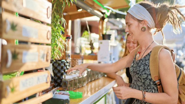 Two women look at jars on a counter at an outdoor market. One woman is reaching for a jar, smiling, while the other stands beside her. There are wooden shelves with items in the background.