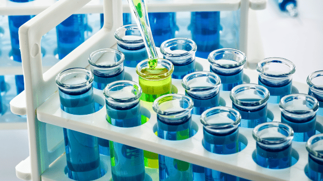 A pipette transferring green liquid into a test tube, surrounded by other test tubes filled with blue liquid, in a laboratory setup.