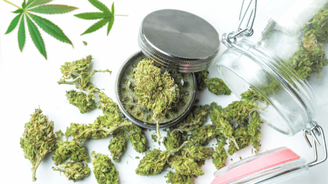 A glass jar with its lid off, spilling cannabis buds onto a white surface. A metal grinder filled with cannabis is nearby, with a cannabis leaf image in the background.