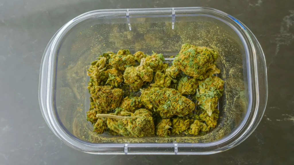 A transparent plastic container with a hinged lid is filled with several cannabis buds. The green buds are dense, coated with visible trichomes, and surrounded by loose fragments and fine particles of the plant. The container is on a dark, smooth surface.