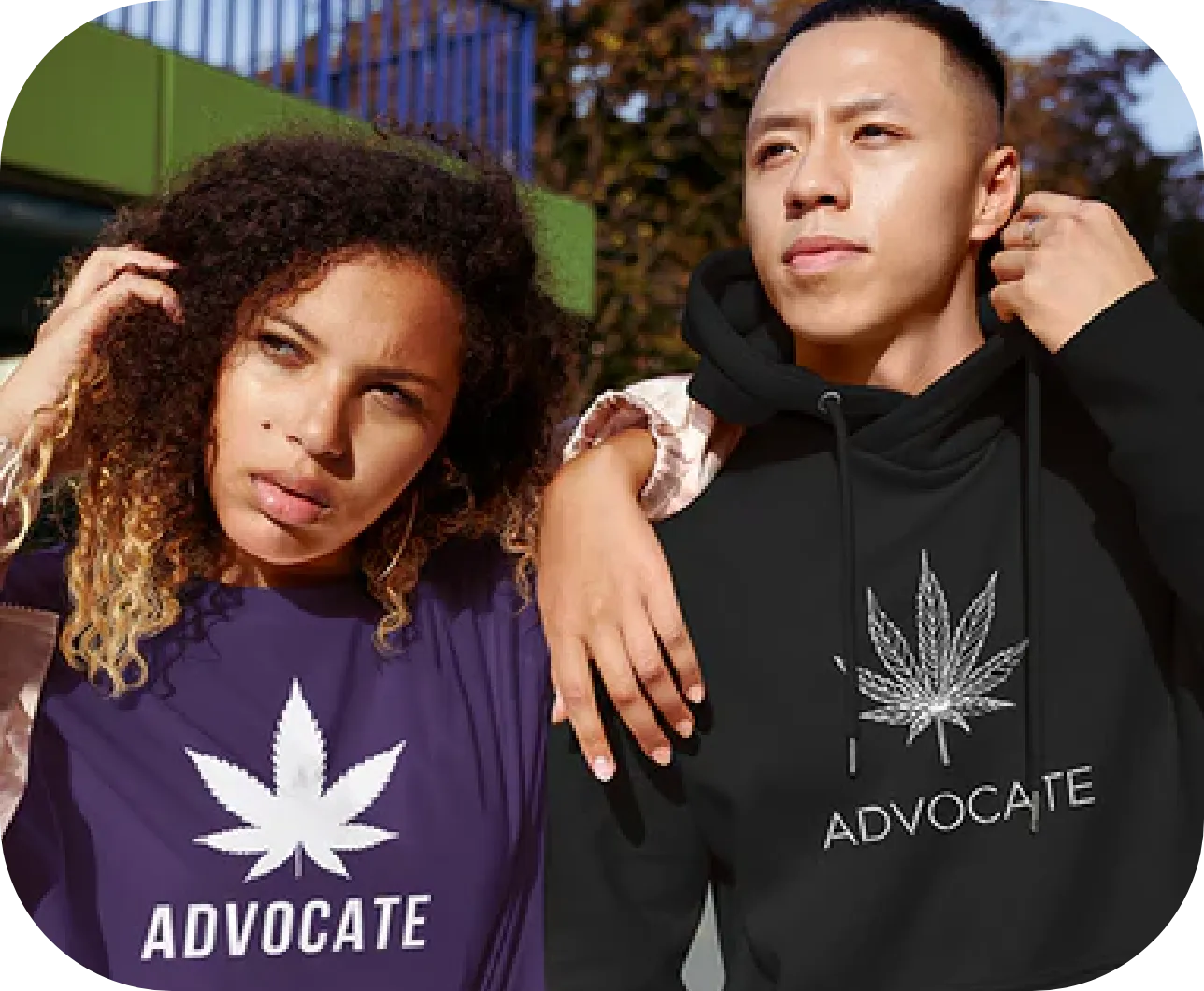 A woman and a man stand side by side, looking confidently into the distance. The woman wears a purple shirt with a white cannabis leaf and the word "ADVOCATE" on it. The man wears a black hoodie with the same design. Trees and a green structure are visible in the background.