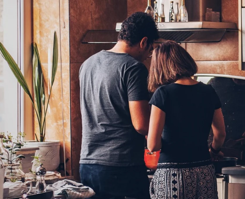 A man and a woman are seen from behind while cooking together in a cozy kitchen. The man is wearing a gray shirt and the woman is in a black top with patterned pants. The kitchen is warmly lit, with potted plants by a large window and various kitchen items on the countertop.