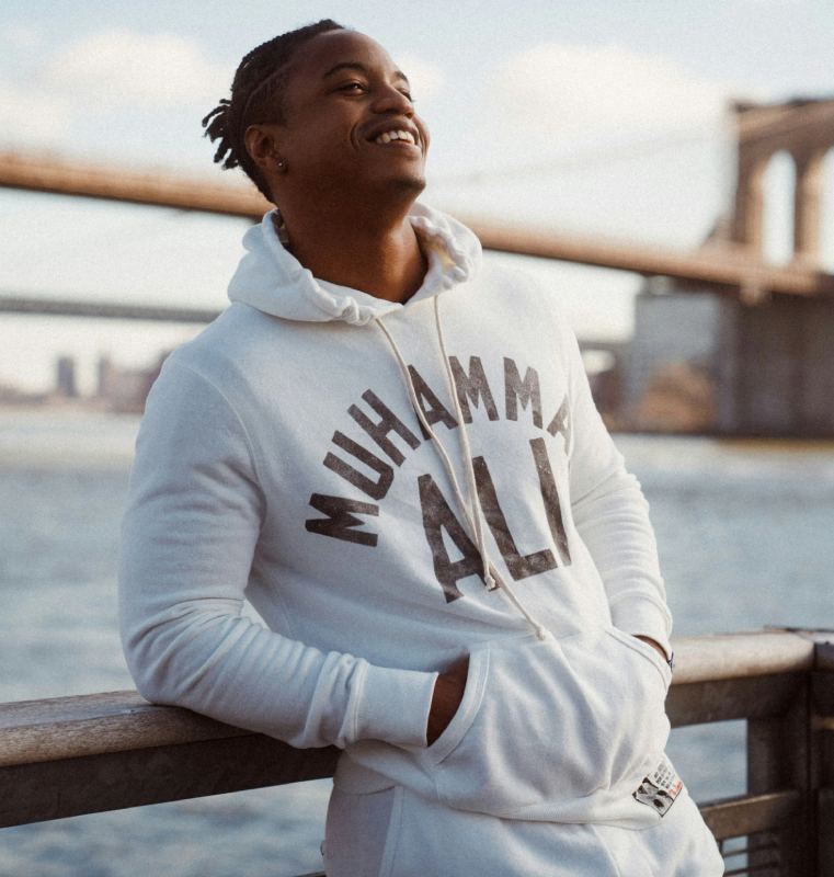 A person stands outdoors by a waterfront, smiling and leaning casually on a railing. They are wearing a white hoodie with the text "MUHAMMAD ALI" on the front. In the background, a bridge extends across the water, and city buildings are faintly visible in the distance.