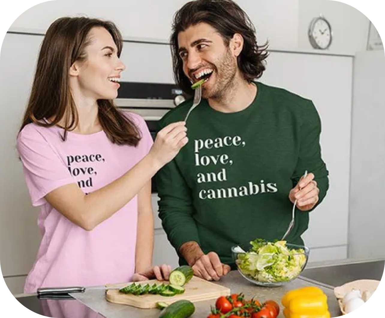 A woman holding a fork feeds a piece of cucumber to a smiling man in a kitchen. The woman wears a pink shirt with the text "peace, love, and" while the man wears a green shirt with the text "peace, love, and cannabis." They are surrounded by fresh vegetables and a bowl of salad.