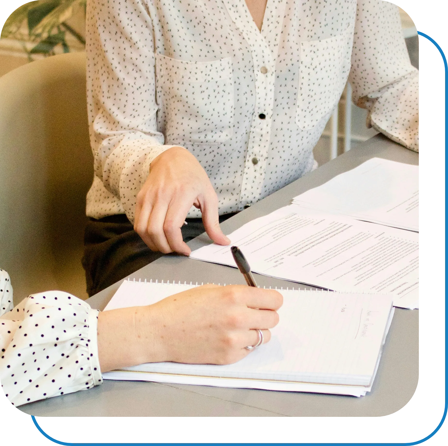 Two individuals are seated at a desk, engaged in paperwork. One person is holding a pen and writing in a notebook, while the other is pointing at a stack of printed documents. Both are wearing light-colored, polka-dotted blouses. The environment suggests a professional or office setting.