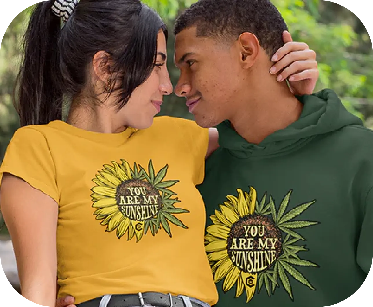 A young couple stands closely, gazing into each other's eyes. The woman wears a yellow T-shirt and the man wears a green hoodie, both featuring a sunflower graphic with the words "YOU ARE MY SUNSHINE" in the center. They are outdoors with blurred greenery in the background.