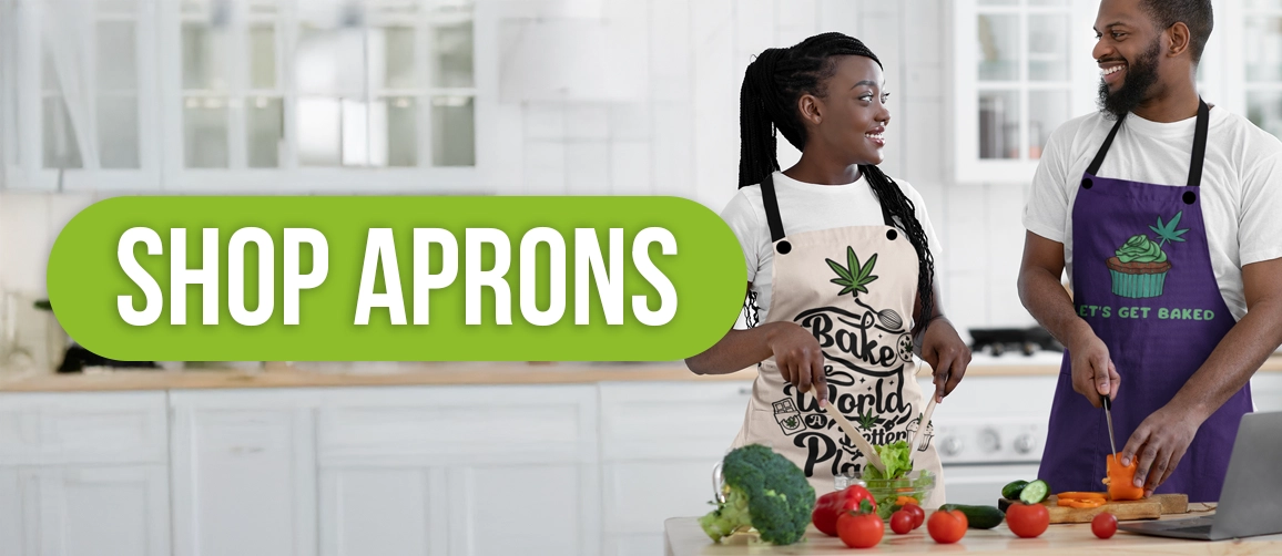 A man and woman in a kitchen are preparing food. Both wear aprons featuring cannabis-themed designs. The woman slices bell peppers, and the man chops carrots. In the background is a countertop with various vegetables. A green banner reads "Shop Aprons.