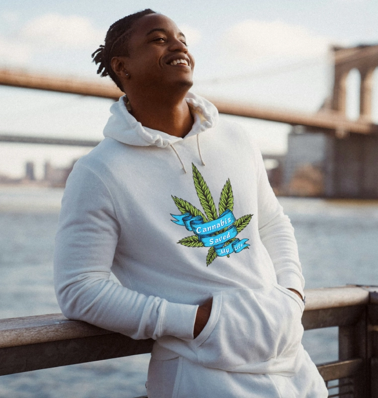 A person wearing a white hoodie with a cannabis leaf design and text "Cannabis Saved My Life" stands smiling on a riverside walkway. Behind them is a large bridge and cityscape under a partly cloudy sky. Their hair is styled in braids, and they lean casually against a metal railing.